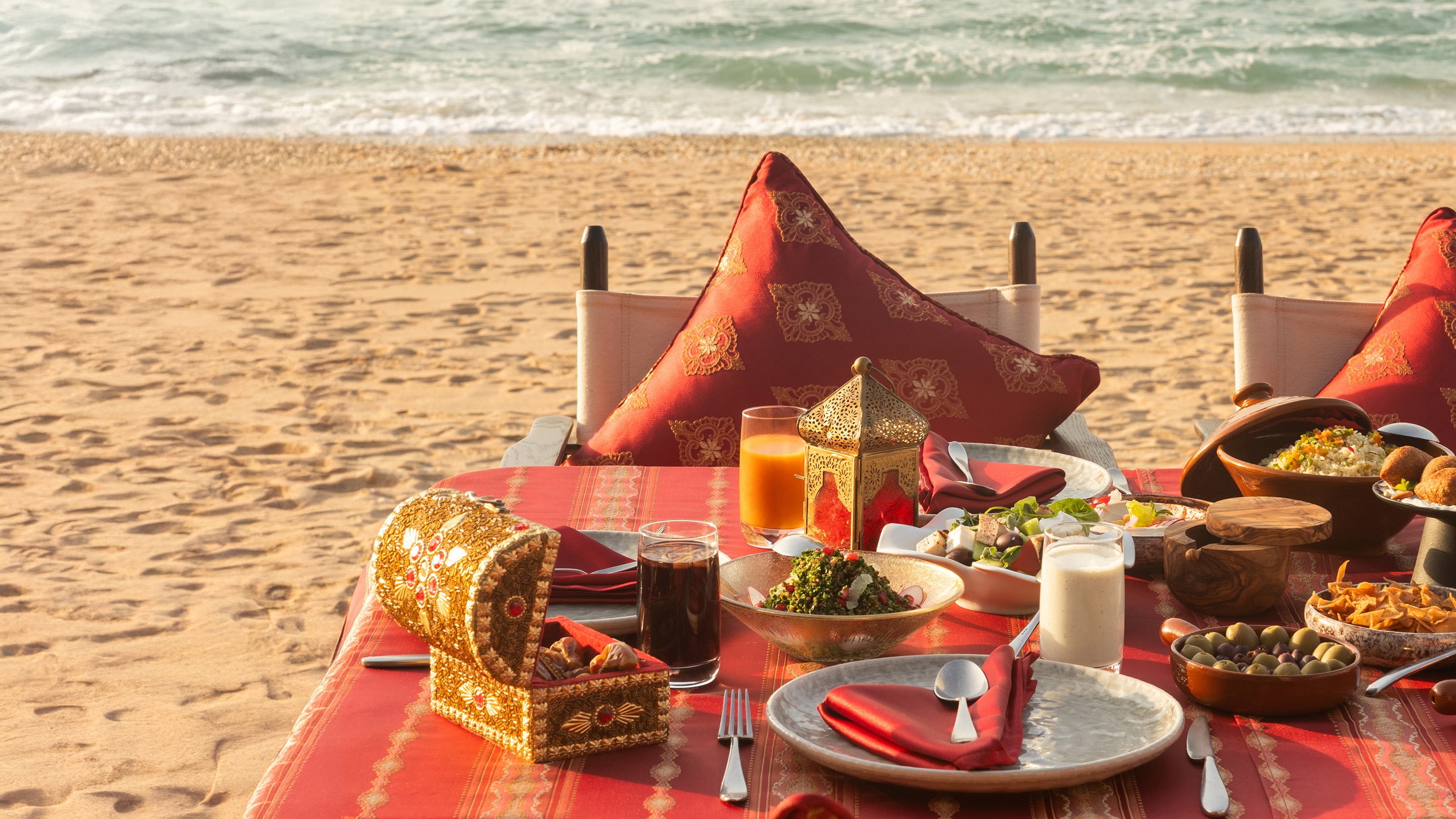 A beautifully set beachside dining table with Middle Eastern dishes, drinks, and decorative cushions overlooking the sea.